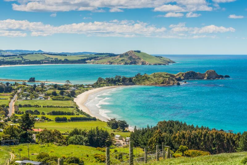 A scenic coastal landscape with turquoise sea, sandy beach, rocky headland, and lush green fields bordered by trees under a partly cloudy blue sky. Houses and farmland are visible near the shoreline.