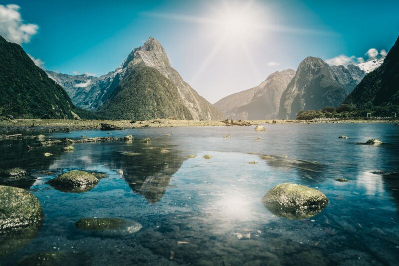 A calm lake with rocks in the foreground reflects tall, jagged mountains under a bright, sunny sky. Sunlight creates a glare on the water and the landscape is lush and green.
