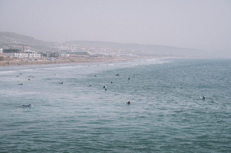 Surfers are scattered across gentle ocean waves near a sandy beach, with distant hills and buildings visible through a hazy sky. The shoreline curves, and people can be seen walking along the beach.