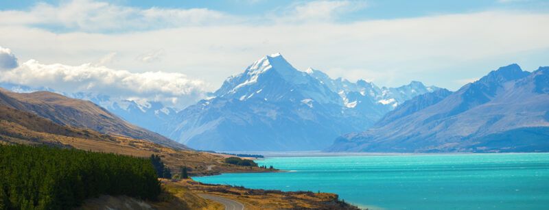 A winding road runs beside a turquoise lake with a backdrop of snow-capped mountains under a partly cloudy blue sky. Grassy hills and dark green trees line the shore, creating a scenic landscape.