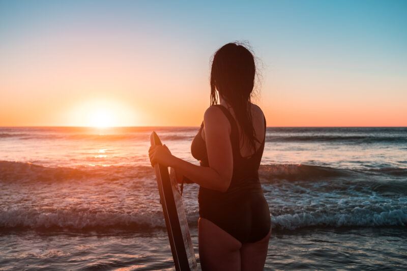 A woman in a black swimsuit holding a surfboard stands on the shore, looking at the ocean during a colorful sunset. Waves are gently rolling in as the sun sets on the horizon.