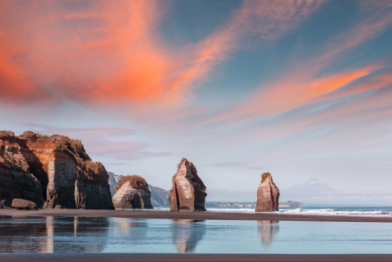 Sea stacks rise from a sandy beach under dramatic orange clouds at sunset, with cliffs on the left and a distant mountain visible on the horizon. The calm water reflects the sky and rock formations.
