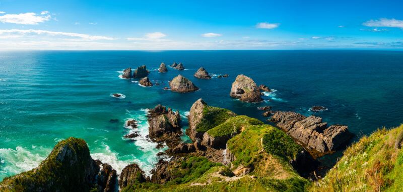 Rocky sea stacks covered in green vegetation rise from vivid blue ocean water under a clear, bright sky, viewed from a grassy coastal cliff. Waves crash around the rocks, creating white foam.