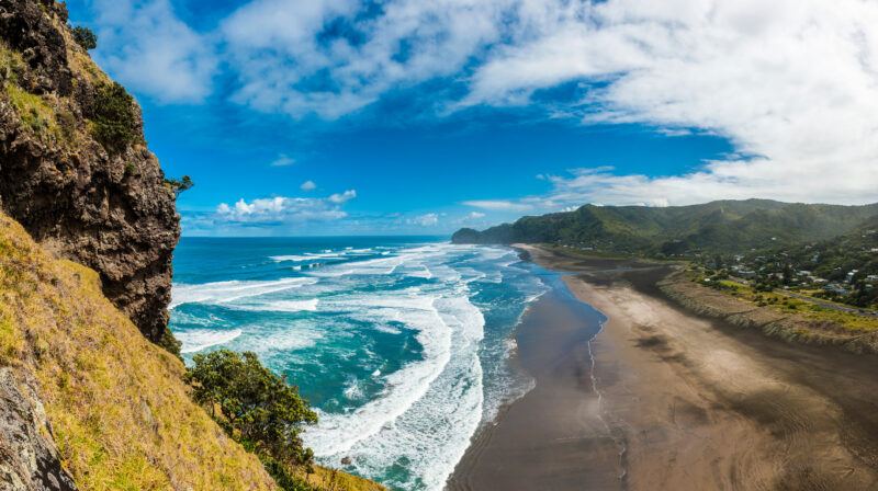 A panoramic view of a coastal landscape with waves crashing onto a wide sandy beach, steep green cliffs on the left, and scattered houses along the shoreline under a partly cloudy blue sky.